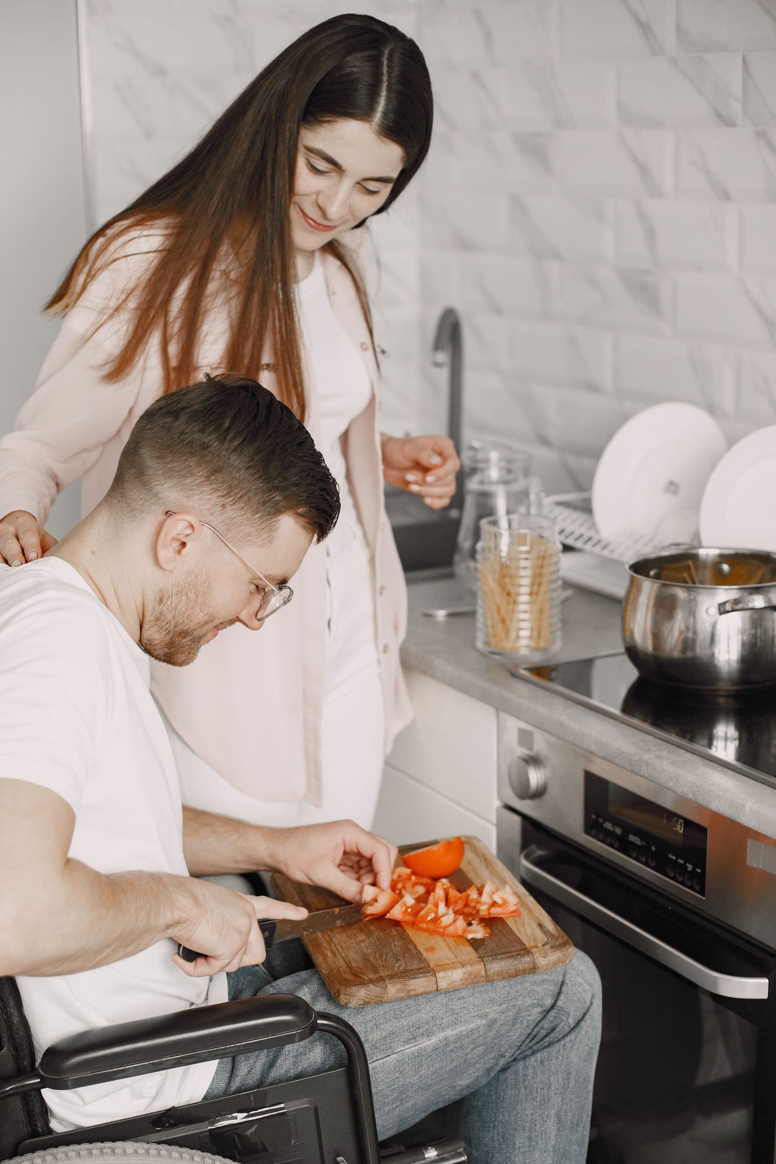 Rapaz em cadeira de rodas corta tomates na cozinha enquanto uma mulher, de pé ao lado dele, acompanha sorrindo.