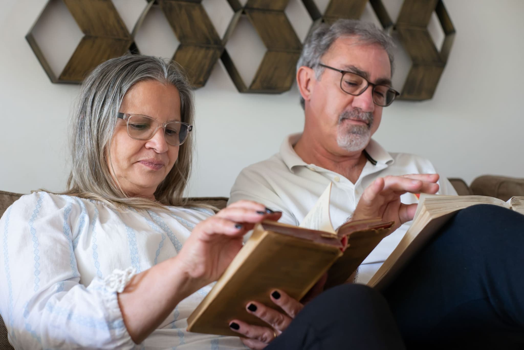 Casal de cabelos grisalhos lê livros lado a lado no sofá de casa.