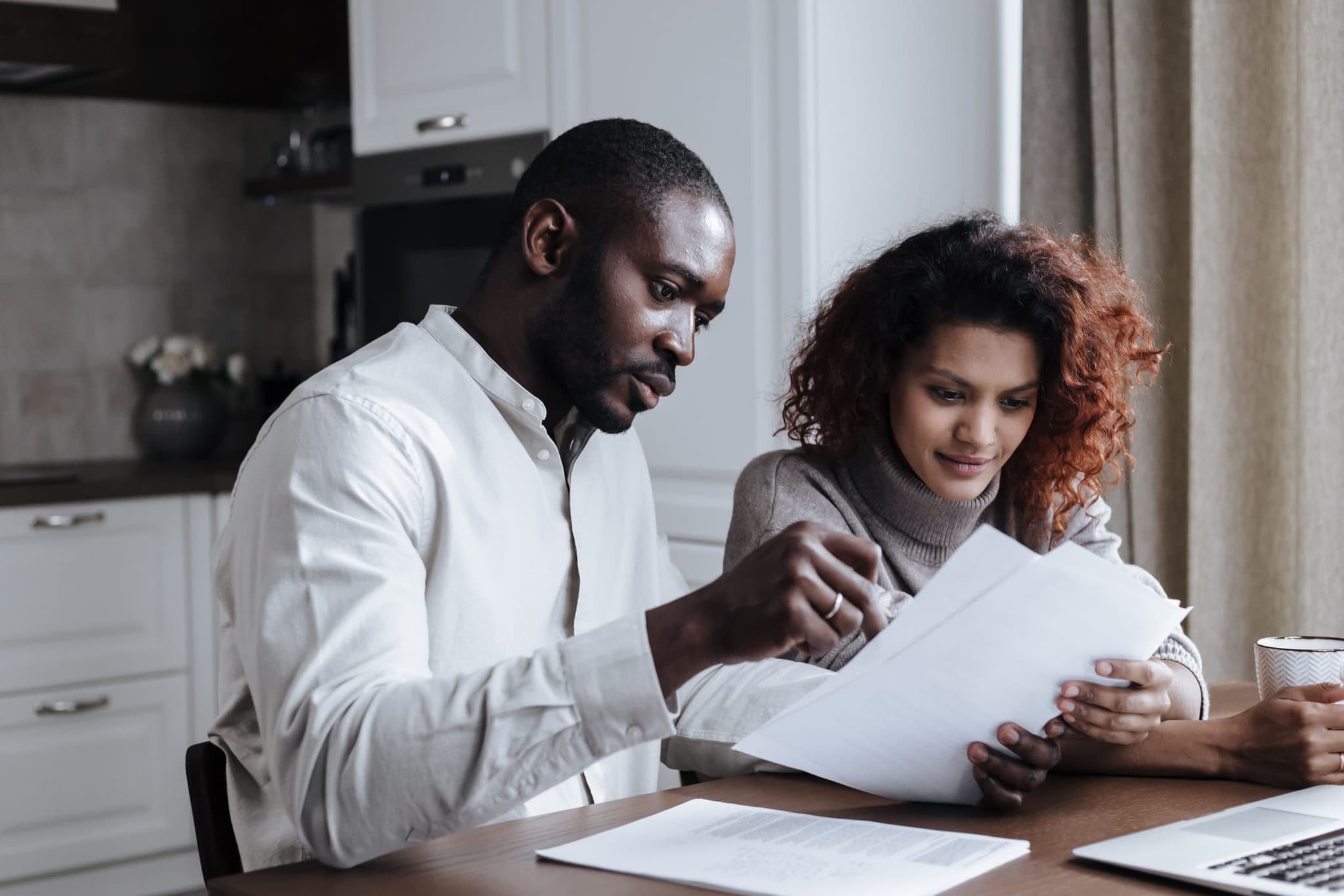 Um homem e uma mulher leem papéis juntos na mesa da cozinha, com um notebook aberto ao lado.