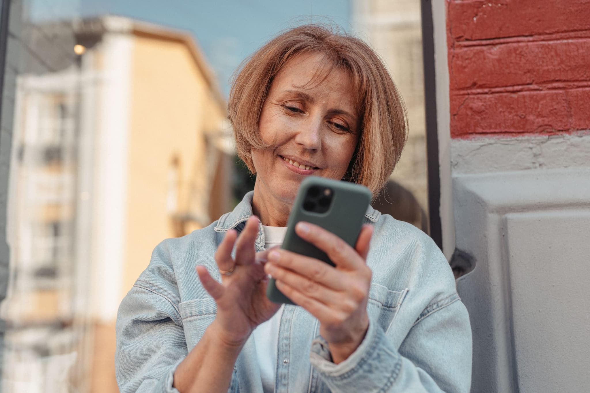 Mulher de uns 55 anos, de jaqueta jeans, sorri enquanto lê uma mensagem no celular, na luz do fim da tarde.