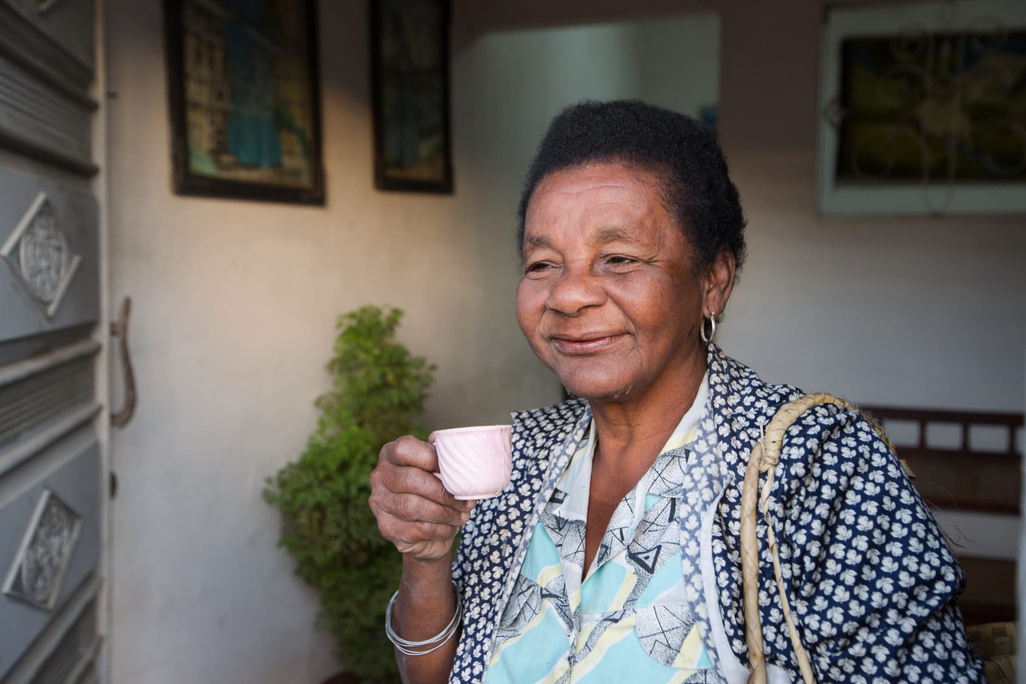 Senhora de cabelo curto sorri tomando um cafezinho numa xícara cor-de-rosa, na entrada de casa.
