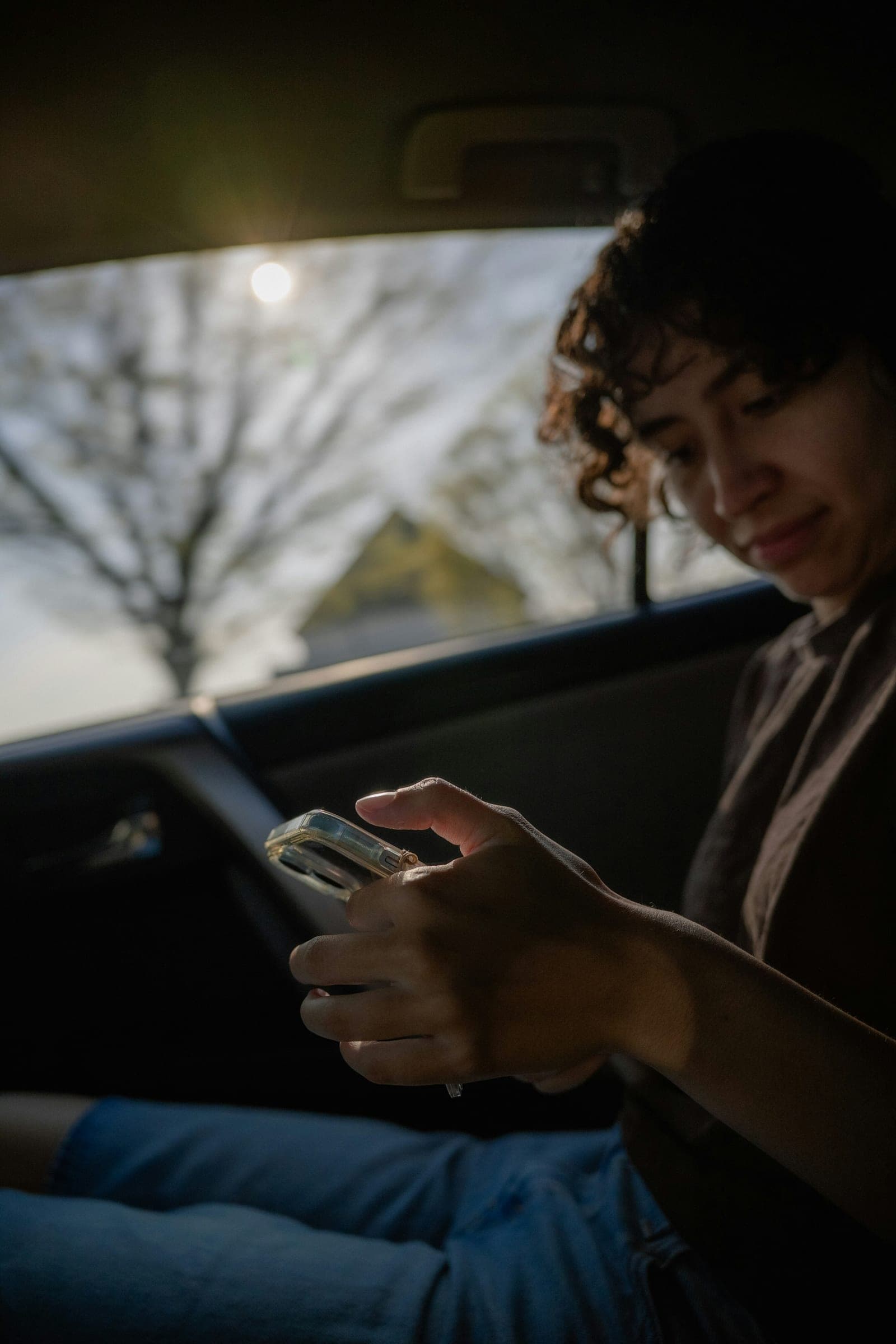 Mulher de cabelo cacheado olha o celular no banco de trás de um carro, com o sol entrando pela janela.