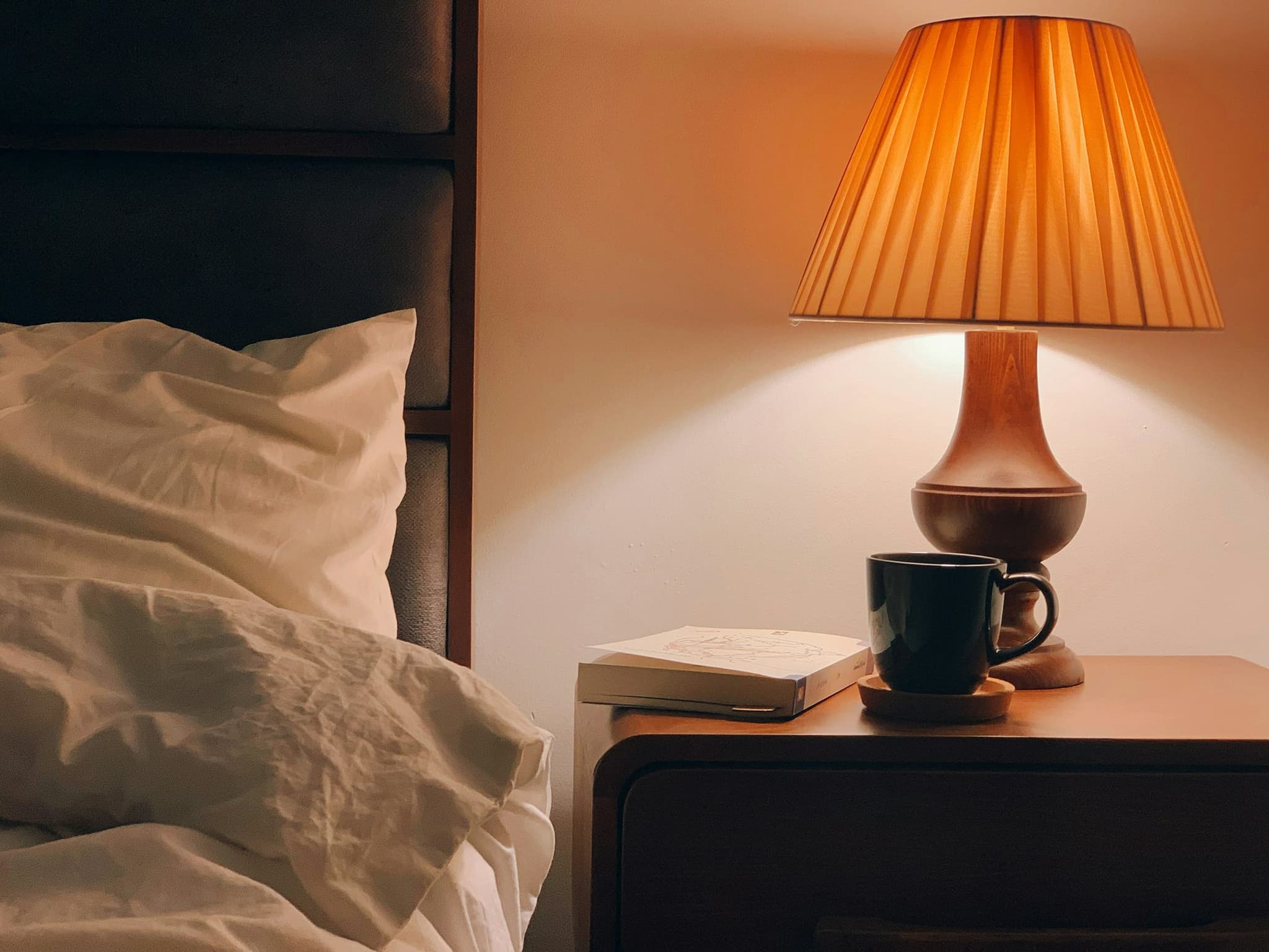 Abajur aceso sobre o criado-mudo, ao lado de uma caneca e de um livro, com a cama desfeita à esquerda.
